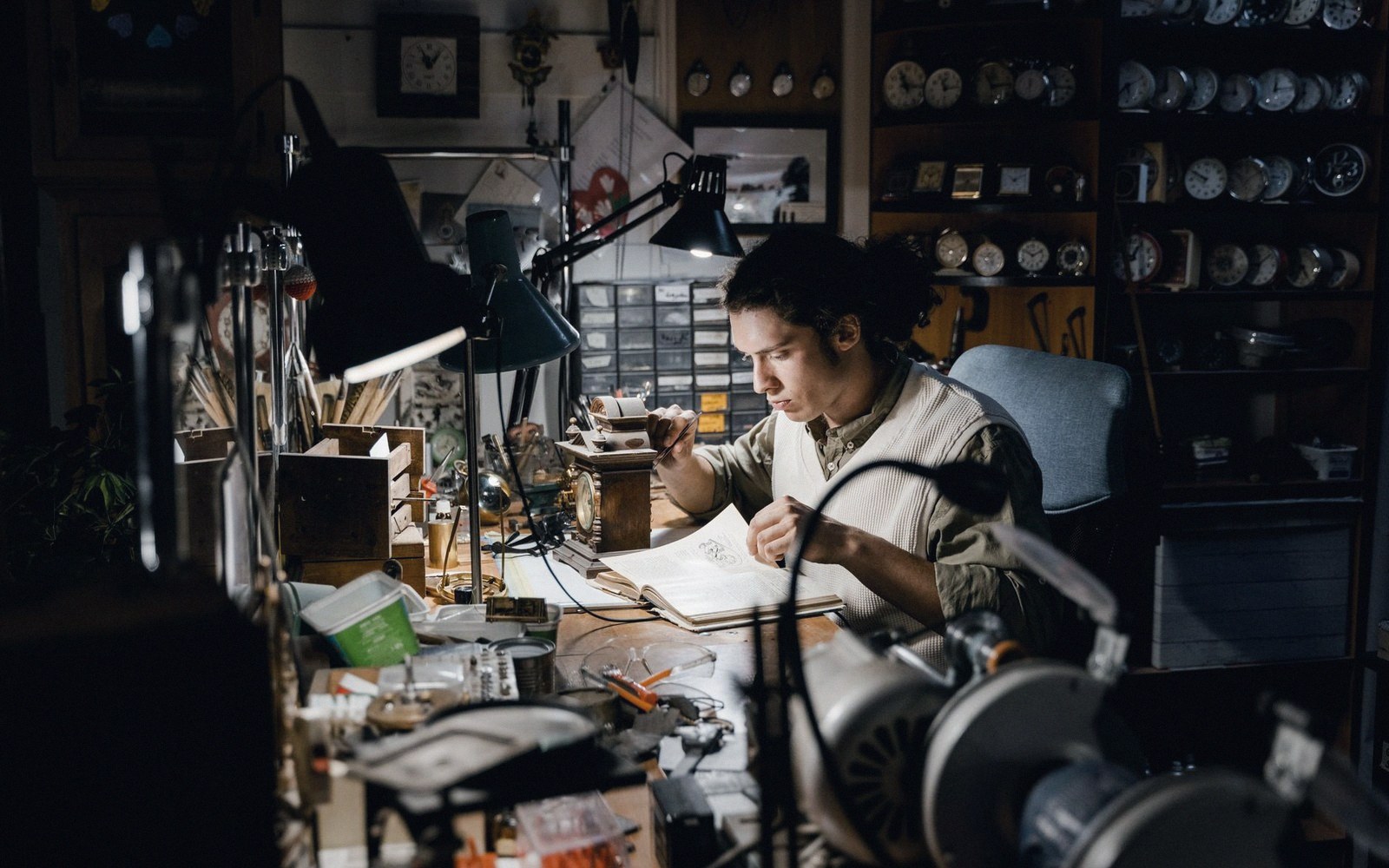 A clockmaker writes in a notebook at a cluttered workbench surrounded by shelves of clocks