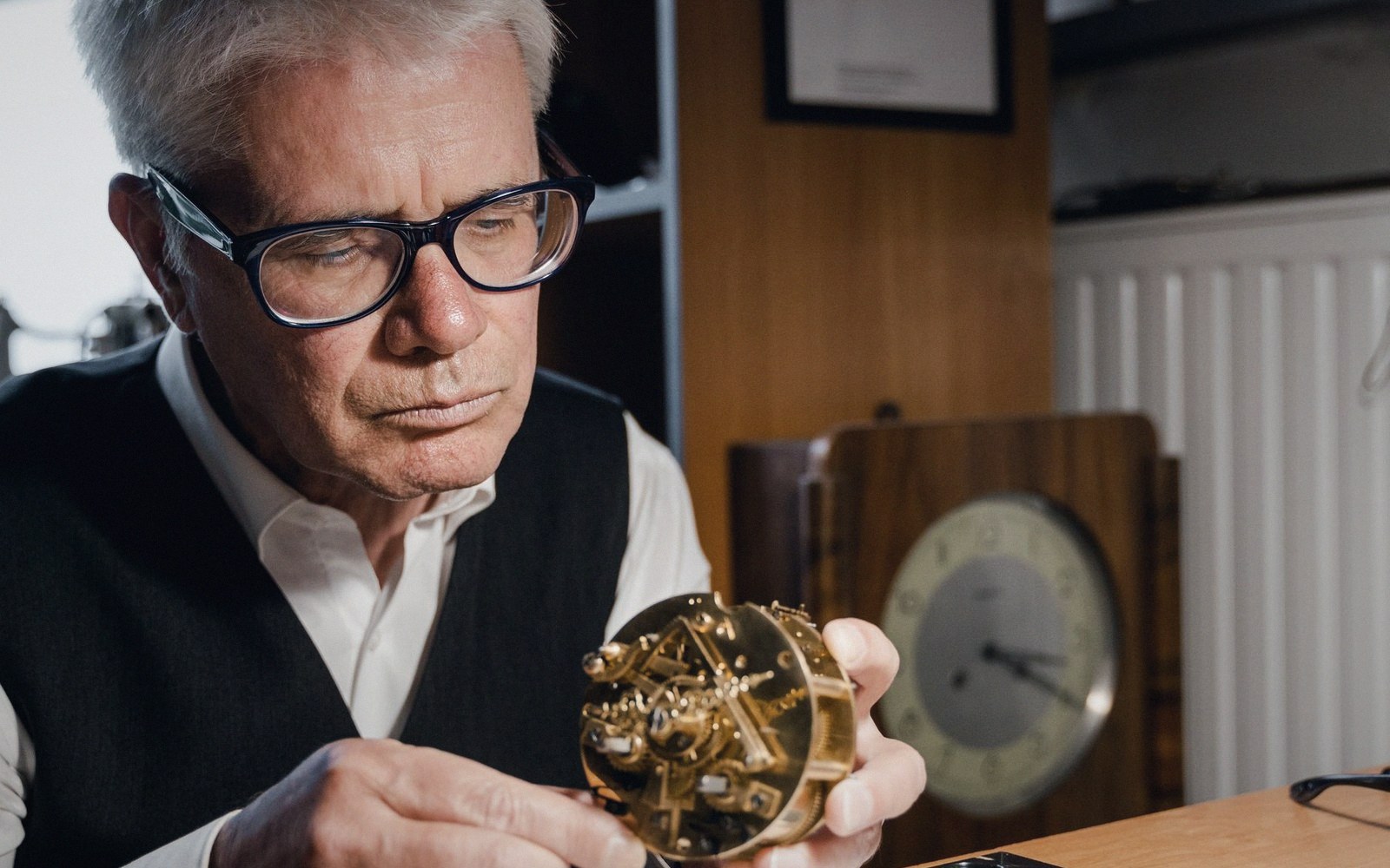 A man in glasses examines an open gold clock movement he holds in his hands