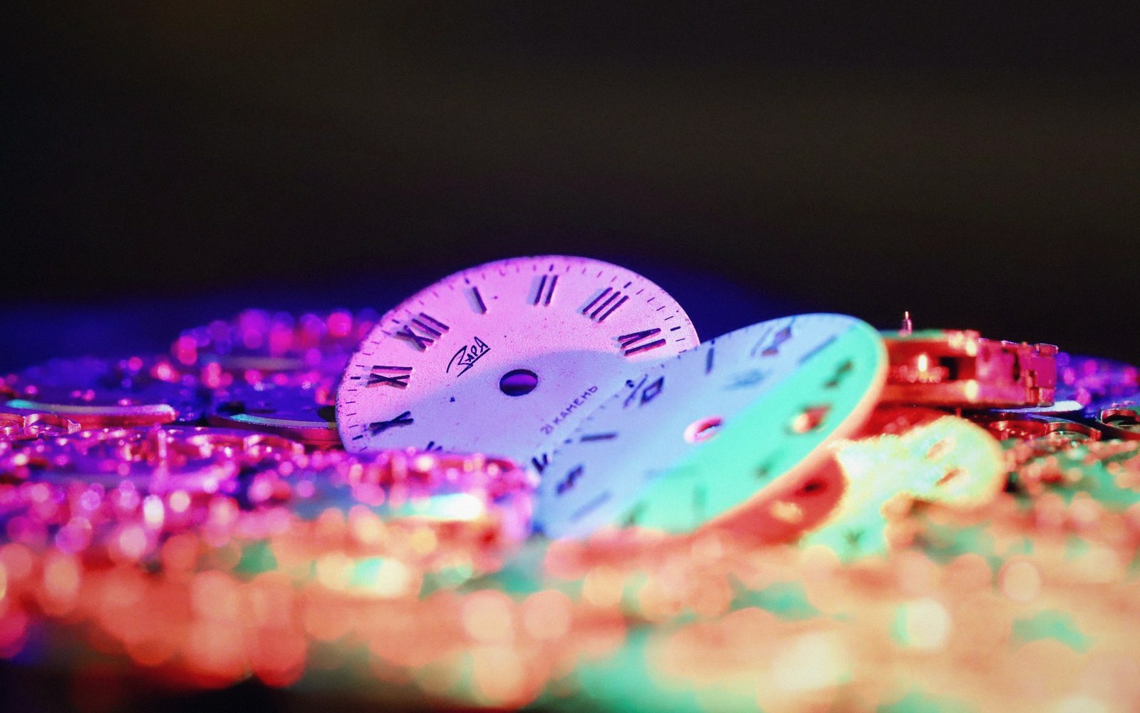 Watch dials with Roman numerals rest among sparkling metal parts under colorful lighting