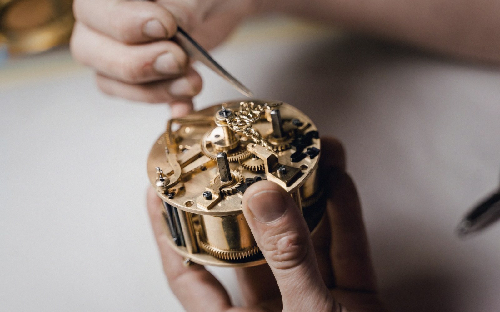 Hands using tweezers to adjust gears inside an exposed brass watch movement